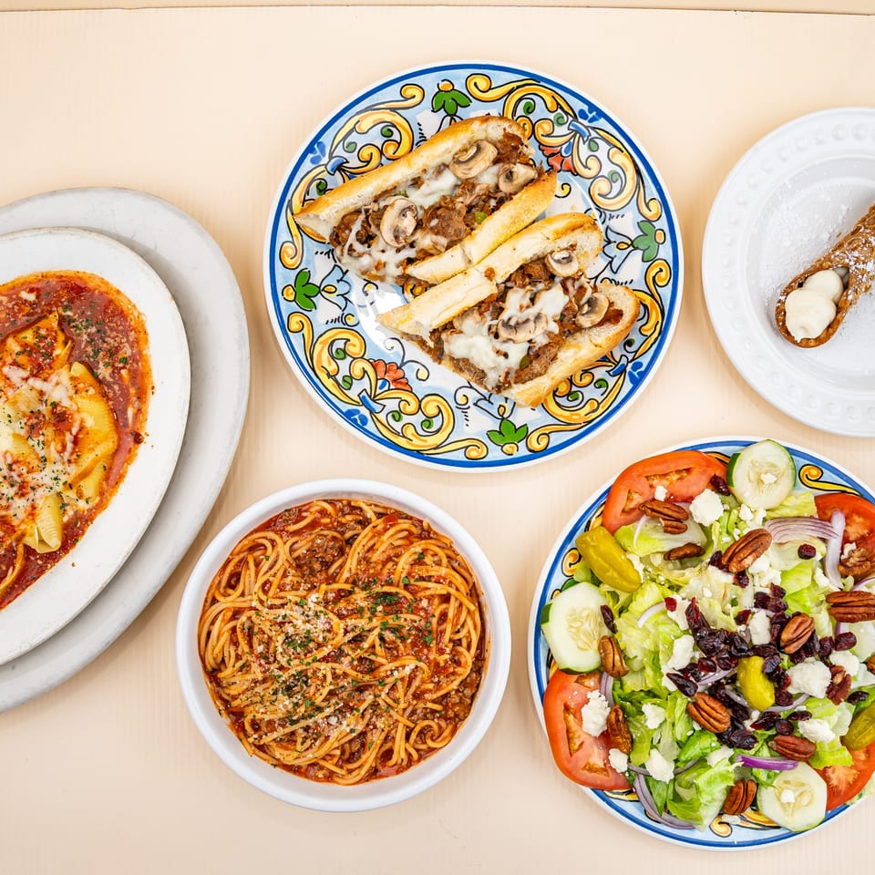 A feast of Italian dishes: baked ziti, spaghetti, cheesesteak subs, a salad, and a cannoli.