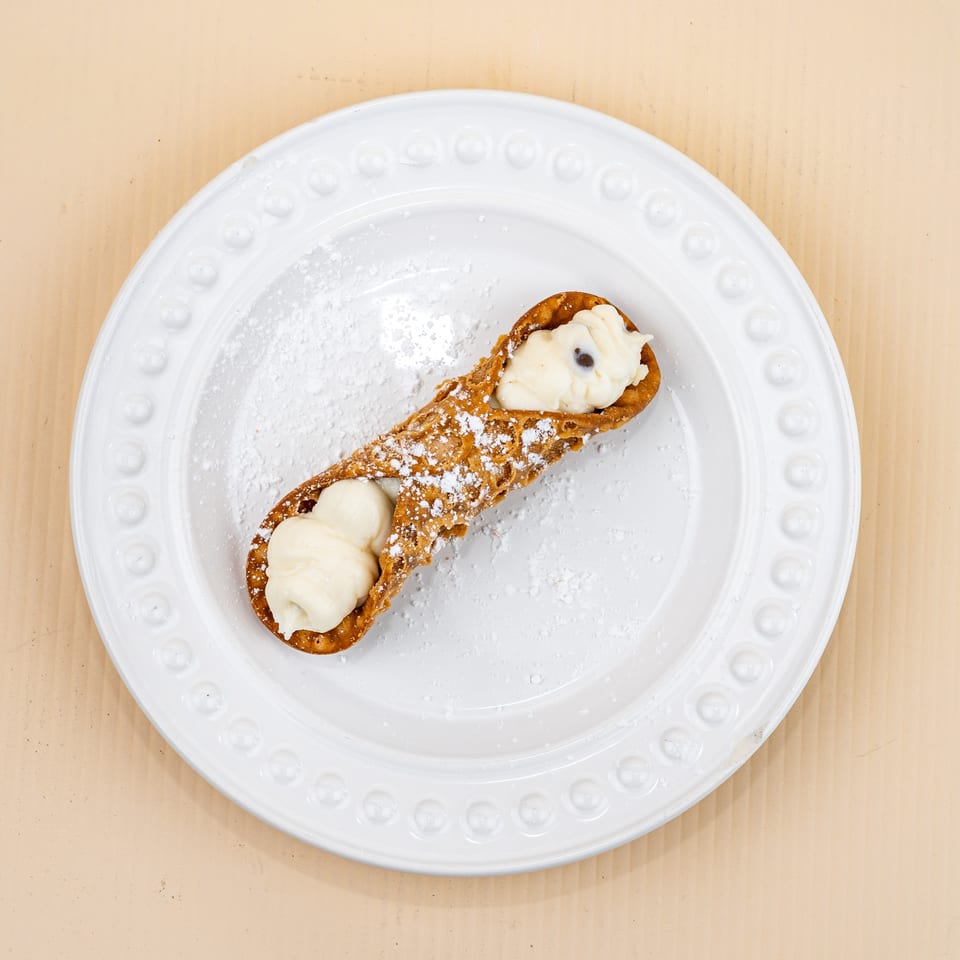 A single cannoli filled with white cream and chocolate chips, dusted with powdered sugar on a white plate.