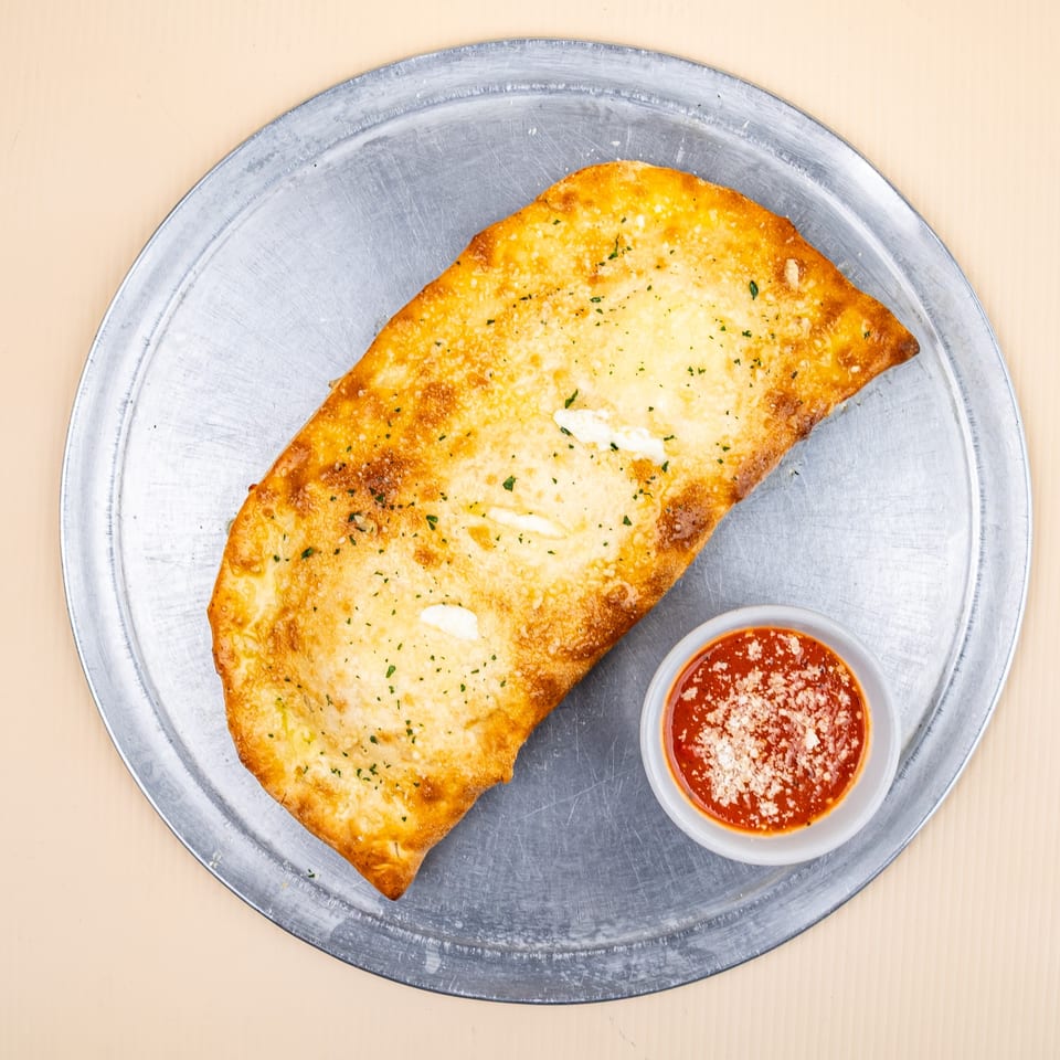 Golden-brown calzone with melted cheese and herbs, served with a side of marinara sauce on a metal tray.